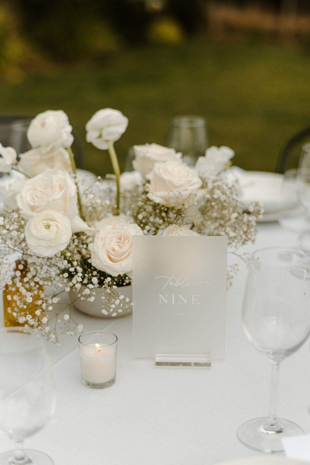 A table decorated with white roses, baby's breath, a "Table Nine" sign, a votive candle, and empty wine glasses set against an outdoor background.