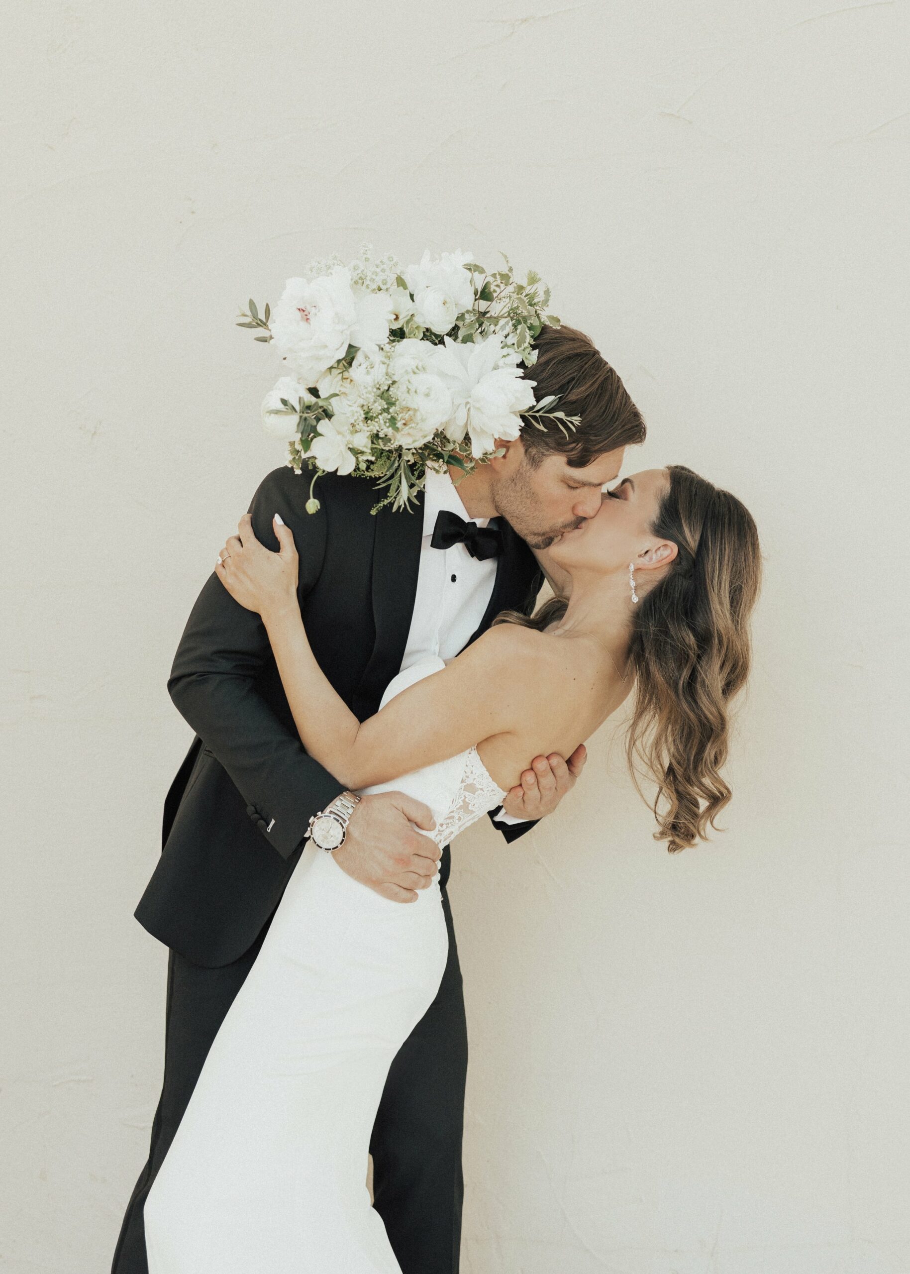 A bride and groom kiss passionately while the groom holds a bouquet of white flowers over their heads. The bride wears a white dress and the groom is in a black suit.