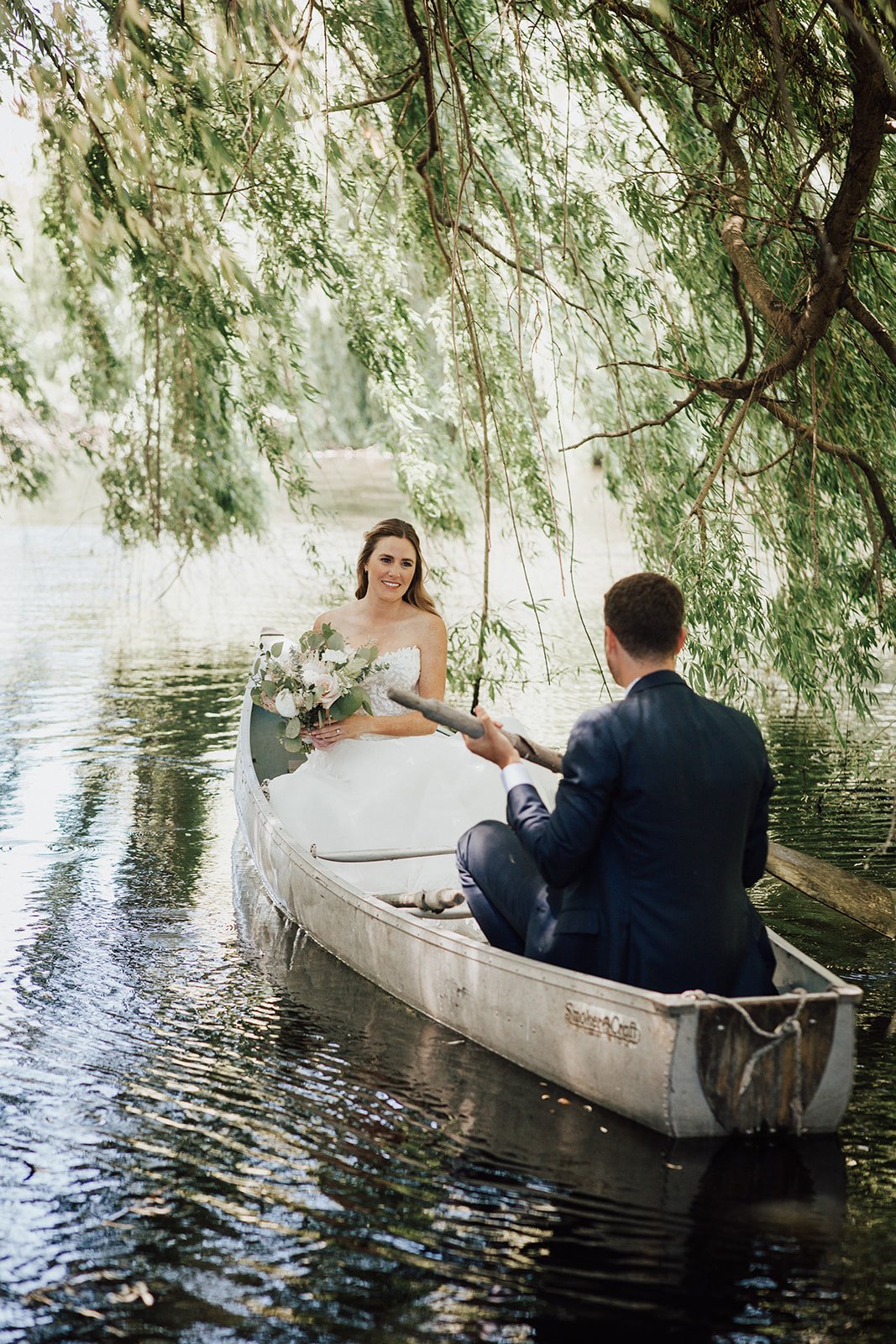 A bride and groom sit in a canoe on a calm, tree-lined river. The groom paddles while the bride holds a bouquet of flowers.