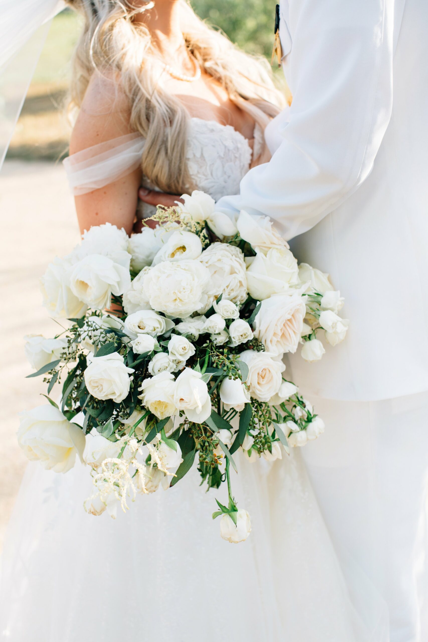 Close-up of a bride holding a bouquet of white roses and greenery, standing with a groom dressed in a white suit.