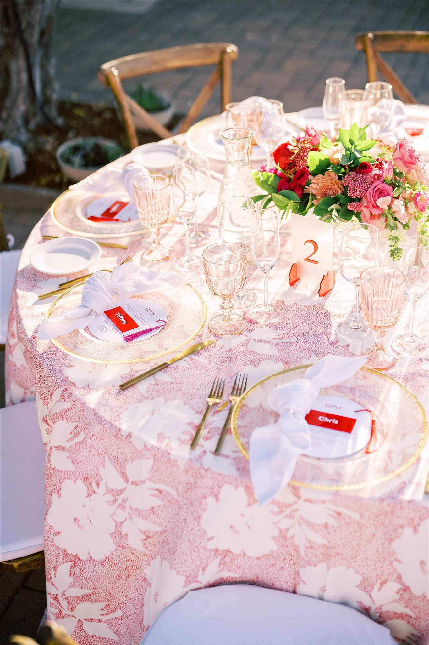 A round table with a floral tablecloth is set for a formal event, featuring plates, glassware, cutlery, and name cards. A floral centerpiece and a table number "2" are also present.