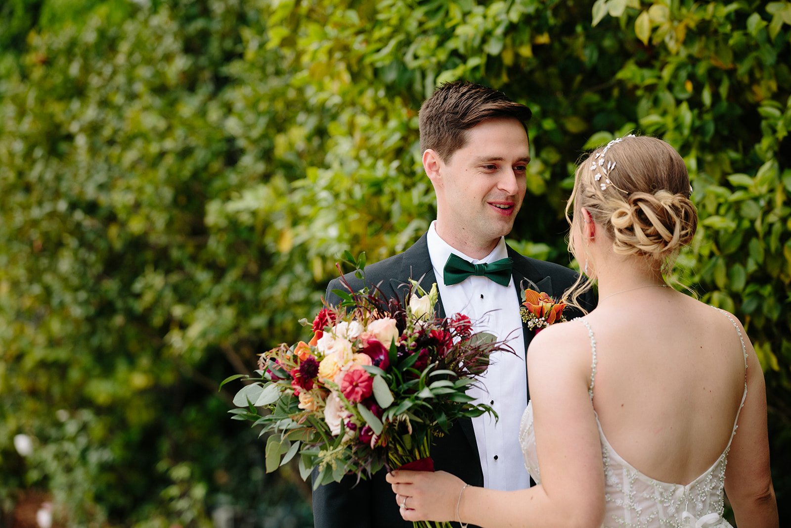 A bride and groom stand facing each other outdoors. The bride holds a bouquet while wearing a white dress with hair in an updo, and the groom wears a dark suit with a green bow tie. Green foliage is in the background.