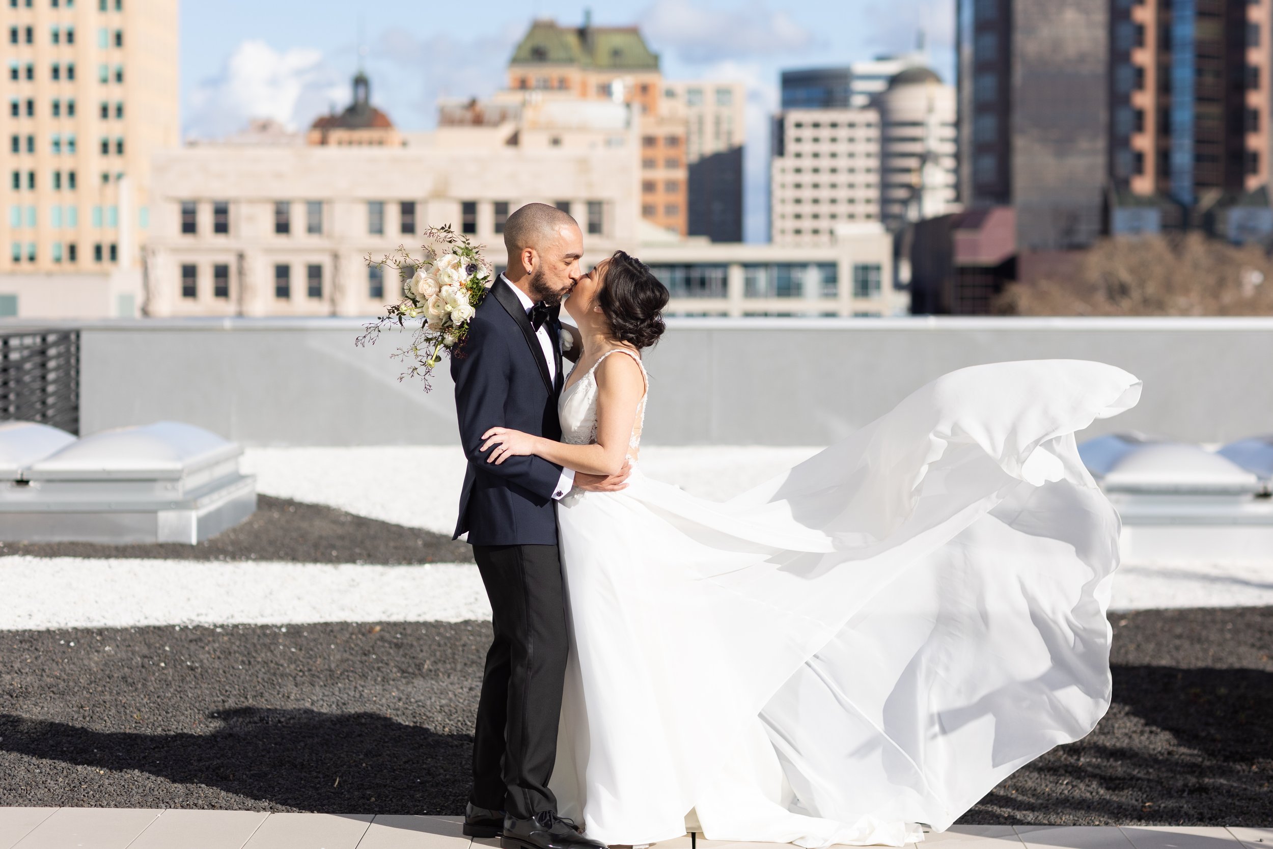 A bride and groom share a kiss on a rooftop, with the bride's dress flowing in the wind and a cityscape in the background.