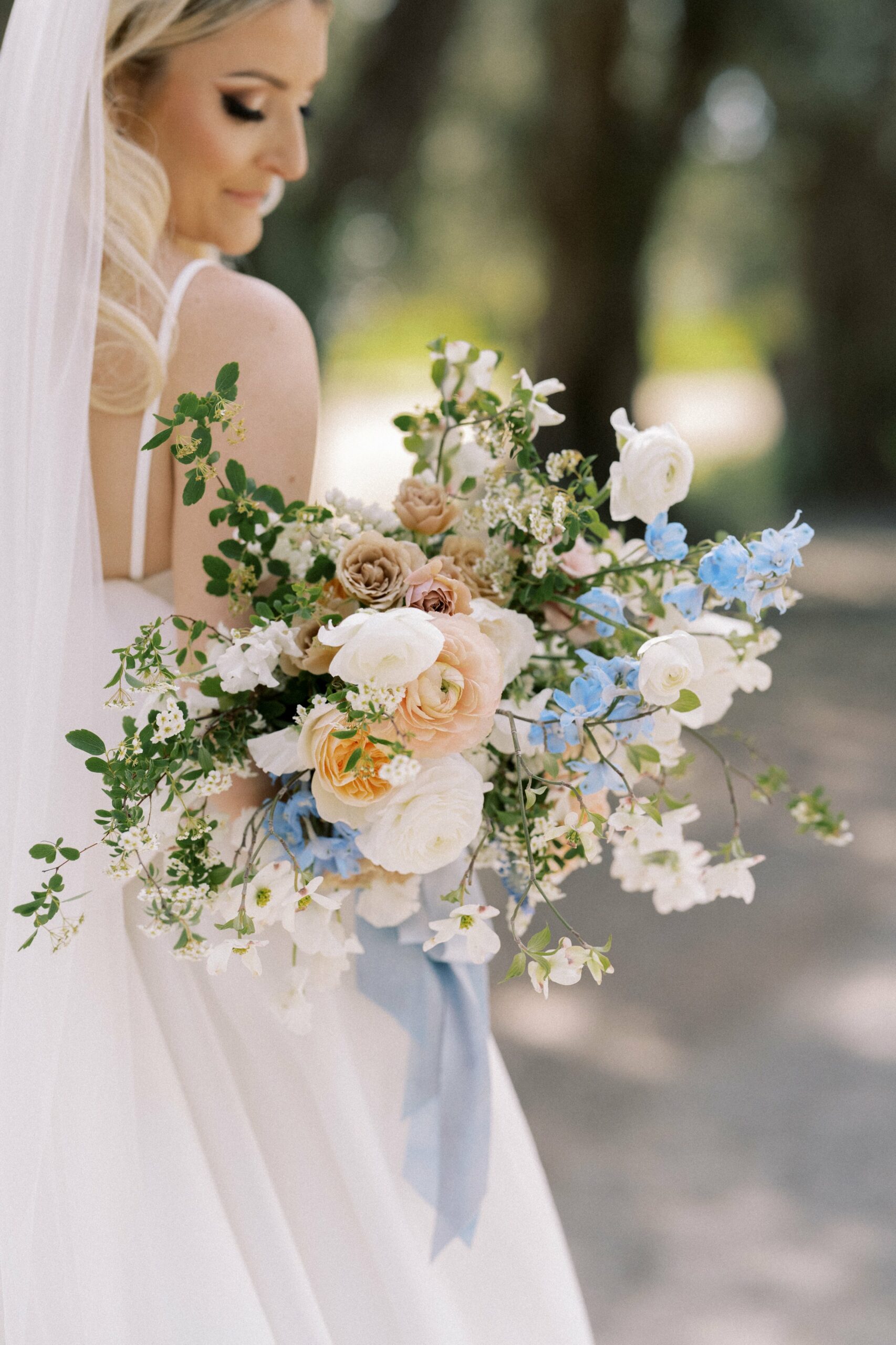 A bride in a sleeveless white dress holds a bouquet of flowers with white, peach, and light blue blooms, standing outdoors with trees in the background.