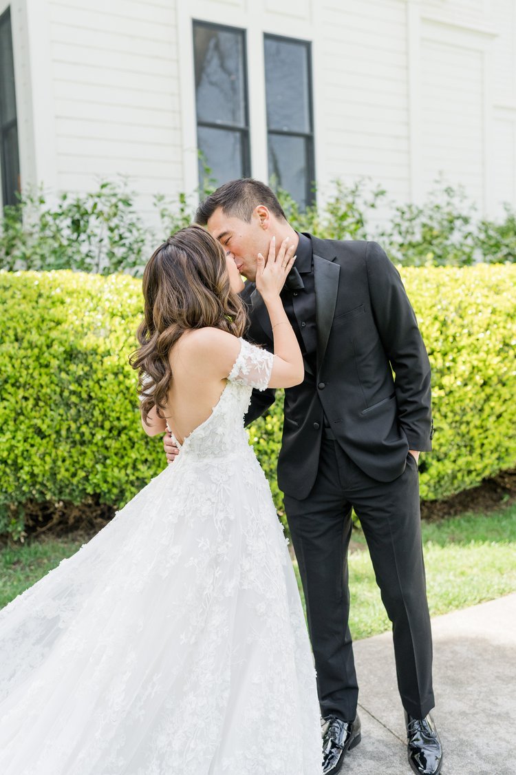 A bride and groom share a kiss outside, standing on a stone path with greenery and a white building in the background. The bride wears a white lace dress, and the groom is dressed in a black suit.