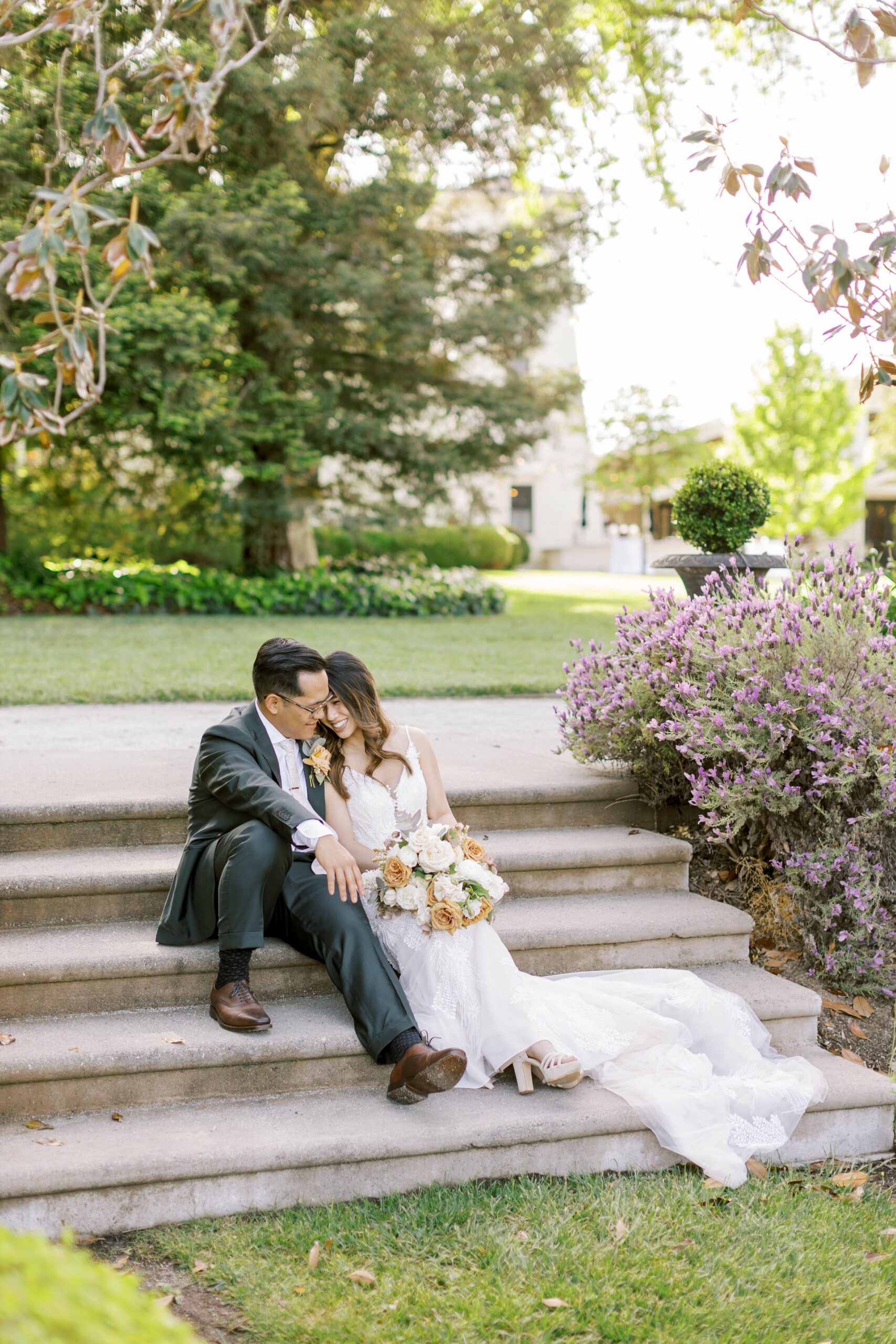 A couple sits on outdoor stone steps, with the woman in a wedding dress holding a bouquet of flowers and the man in a suit leaning affectionately towards her. They are surrounded by greenery and purple shrubs.