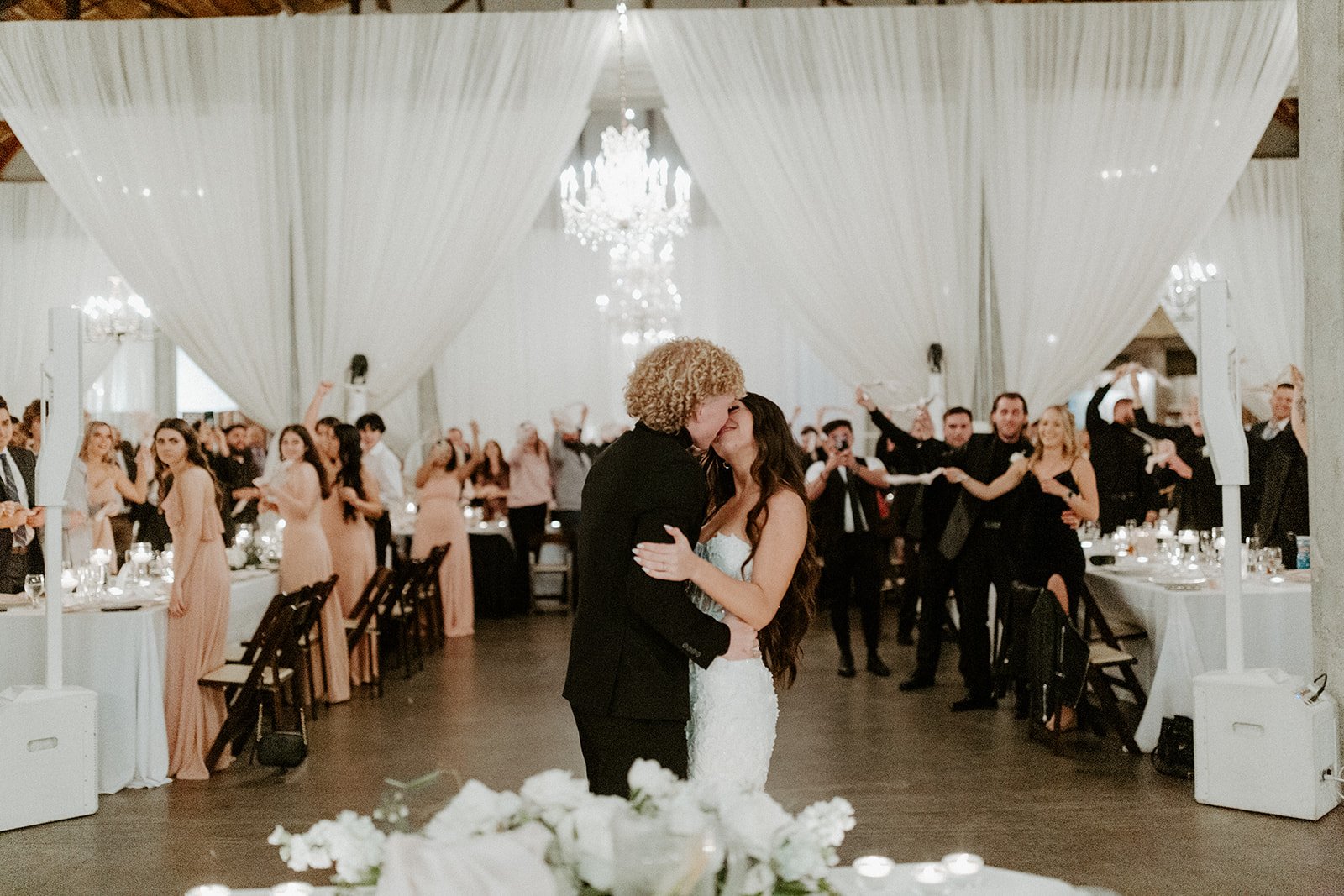 A couple shares a kiss during their wedding reception, surrounded by guests. The venue is elegantly decorated with white drapes and a chandelier. Guests are standing and clapping in the background.