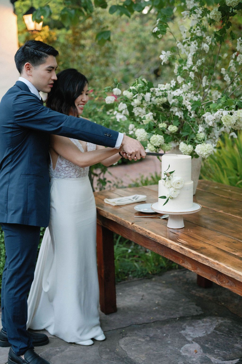 A couple dressed in wedding attire cuts a white-tiered cake on a wooden table adorned with floral arrangements.