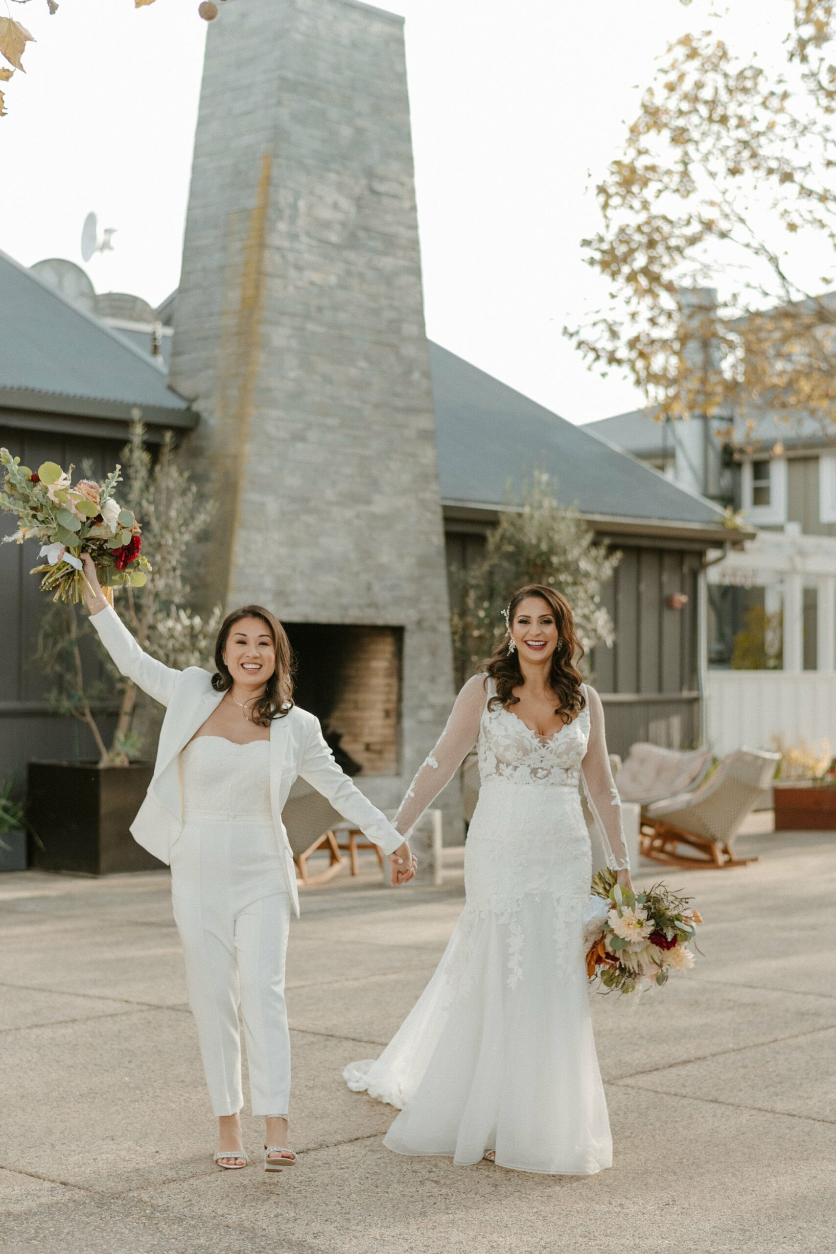 Two women holding hands and smiling, one in a white suit and the other in a white wedding dress, posing outdoors in front of a modern stone structure with bouquets of flowers.