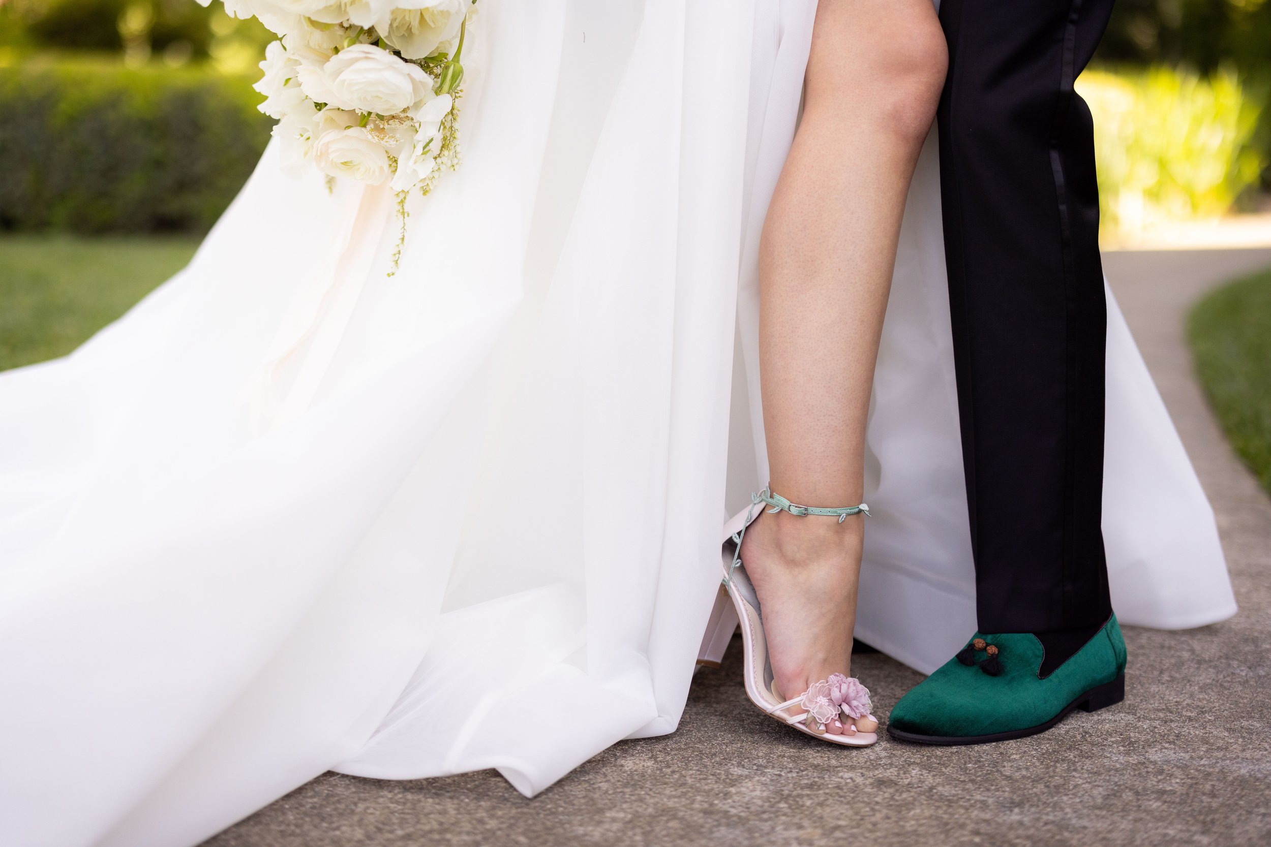 Wedding couple shown from the knees down; the bride in a white gown with white heels and the groom in black pants with green loafers. The bride holds a bouquet of white flowers. Both standing on pavement.