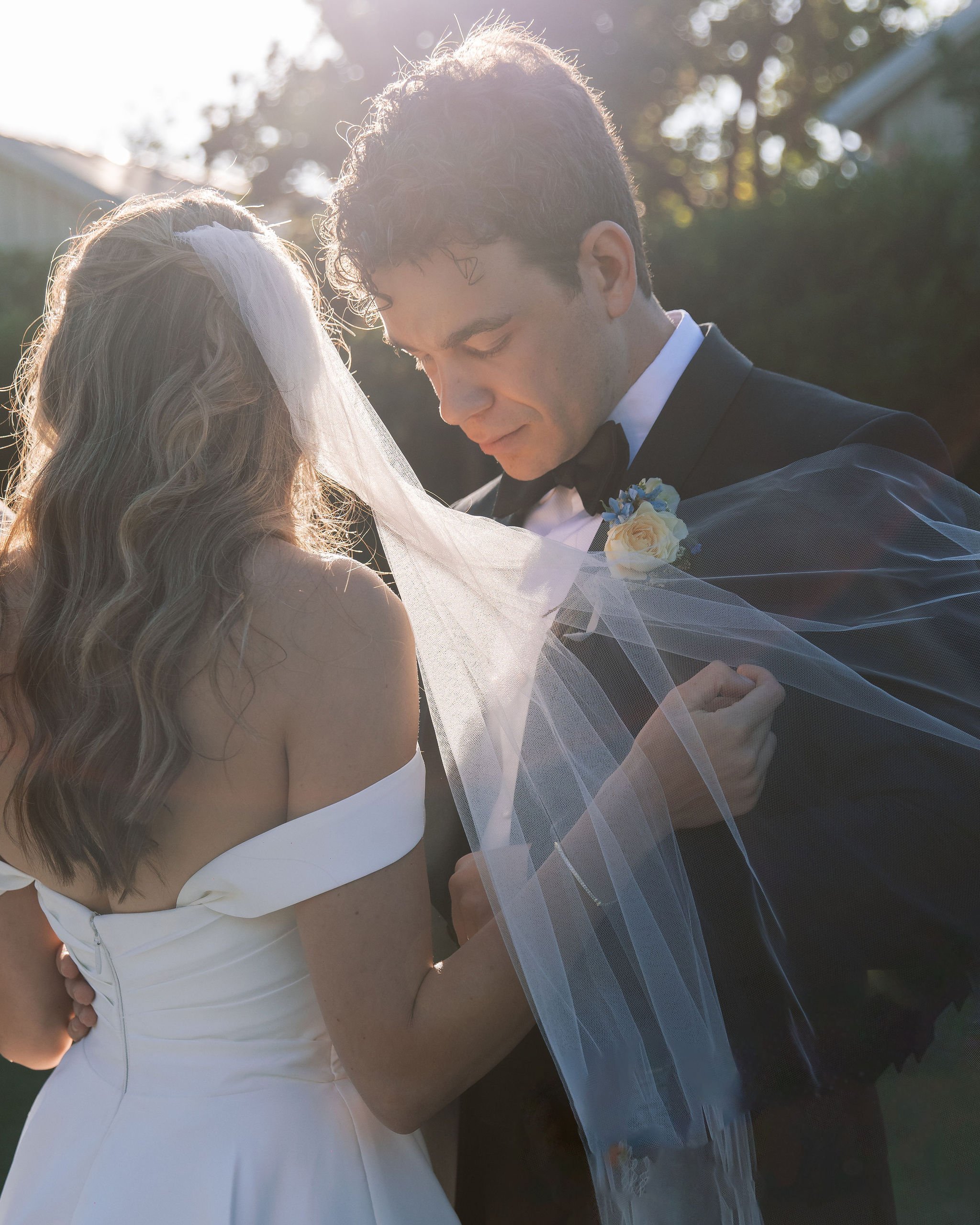 A bride and groom stand closely, with the groom adjusting the bride's veil. They are outdoors, with sunlight filtering through trees in the background.