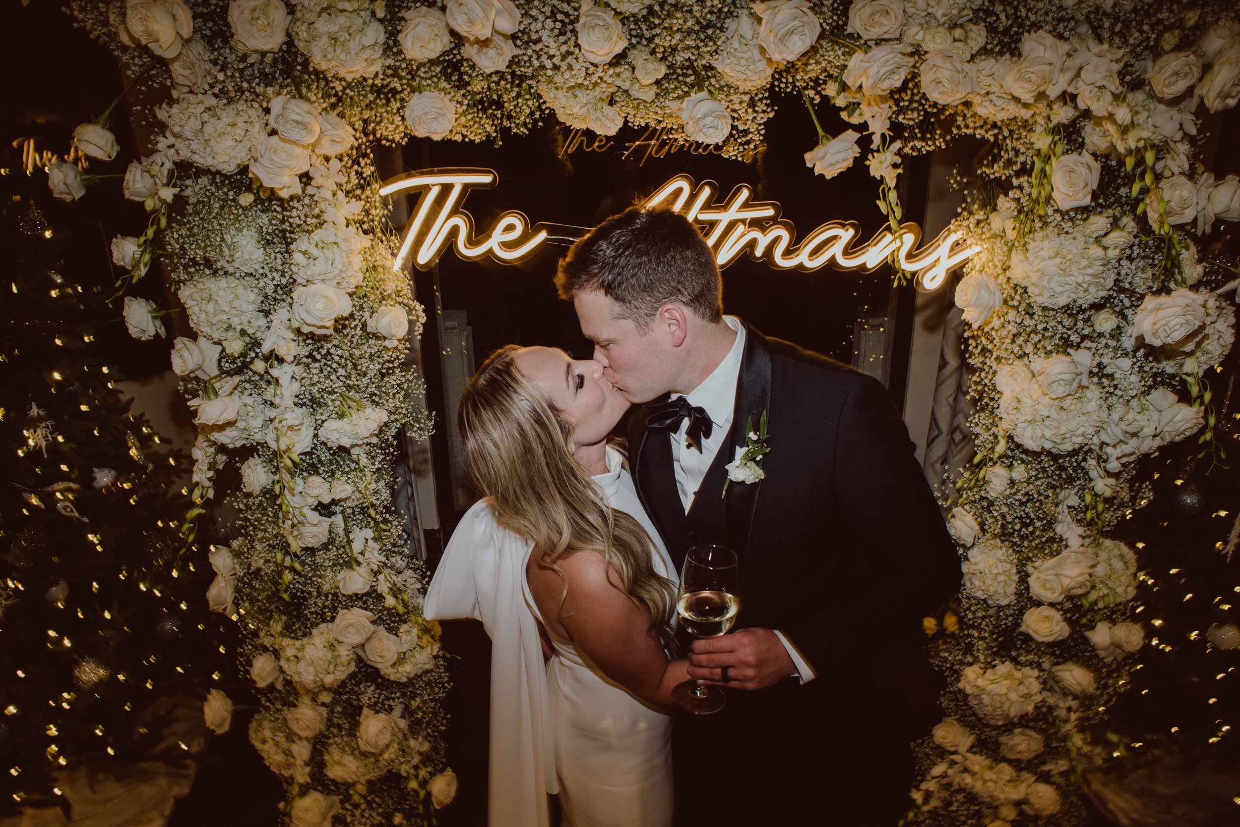 A couple, dressed in wedding attire, shares a kiss under a floral arch with a neon sign in the background. The woman holds a wine glass and wears a white gown, while the man is in a black suit.