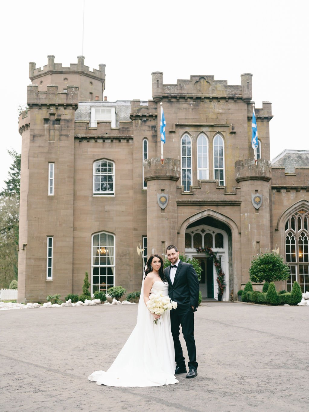 A bride and groom stand outside a large, castle-like building. The bride is wearing a white wedding dress and holding flowers, while the groom is dressed in a black suit with a bow tie.