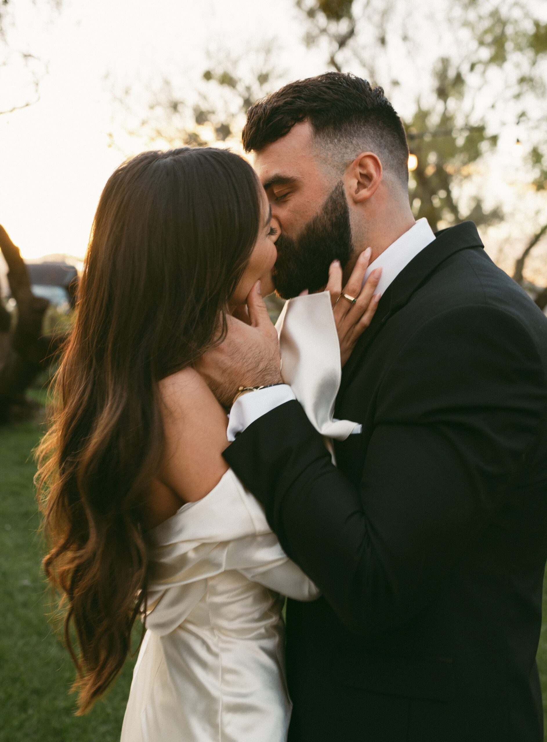 A couple dressed in formal attire shares a kiss outdoors at sunset. The woman is wearing a white dress, and the man is in a black suit.