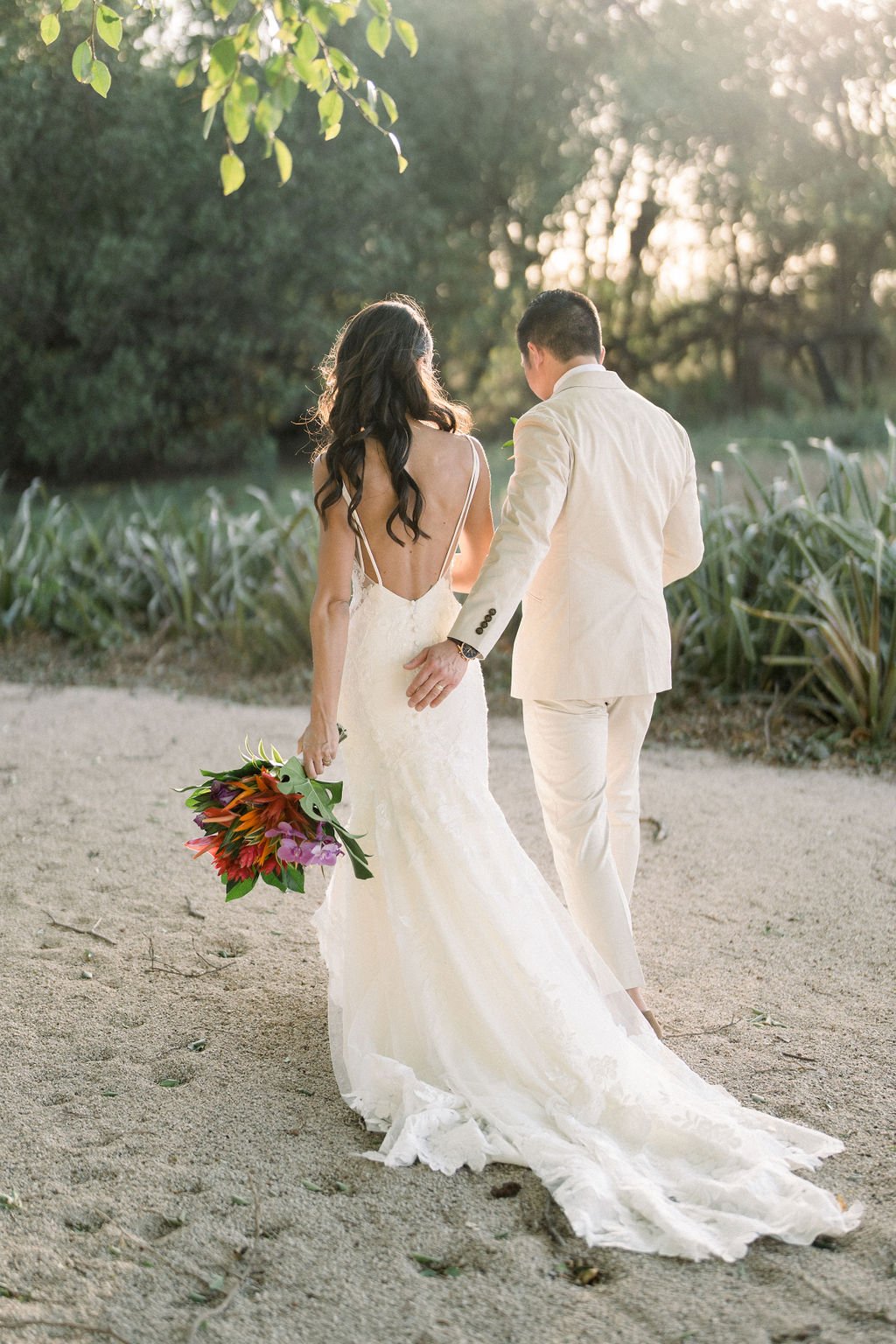 A bride and groom, dressed in white, walk hand in hand on a sandy path, surrounded by greenery. The bride carries a colorful bouquet.