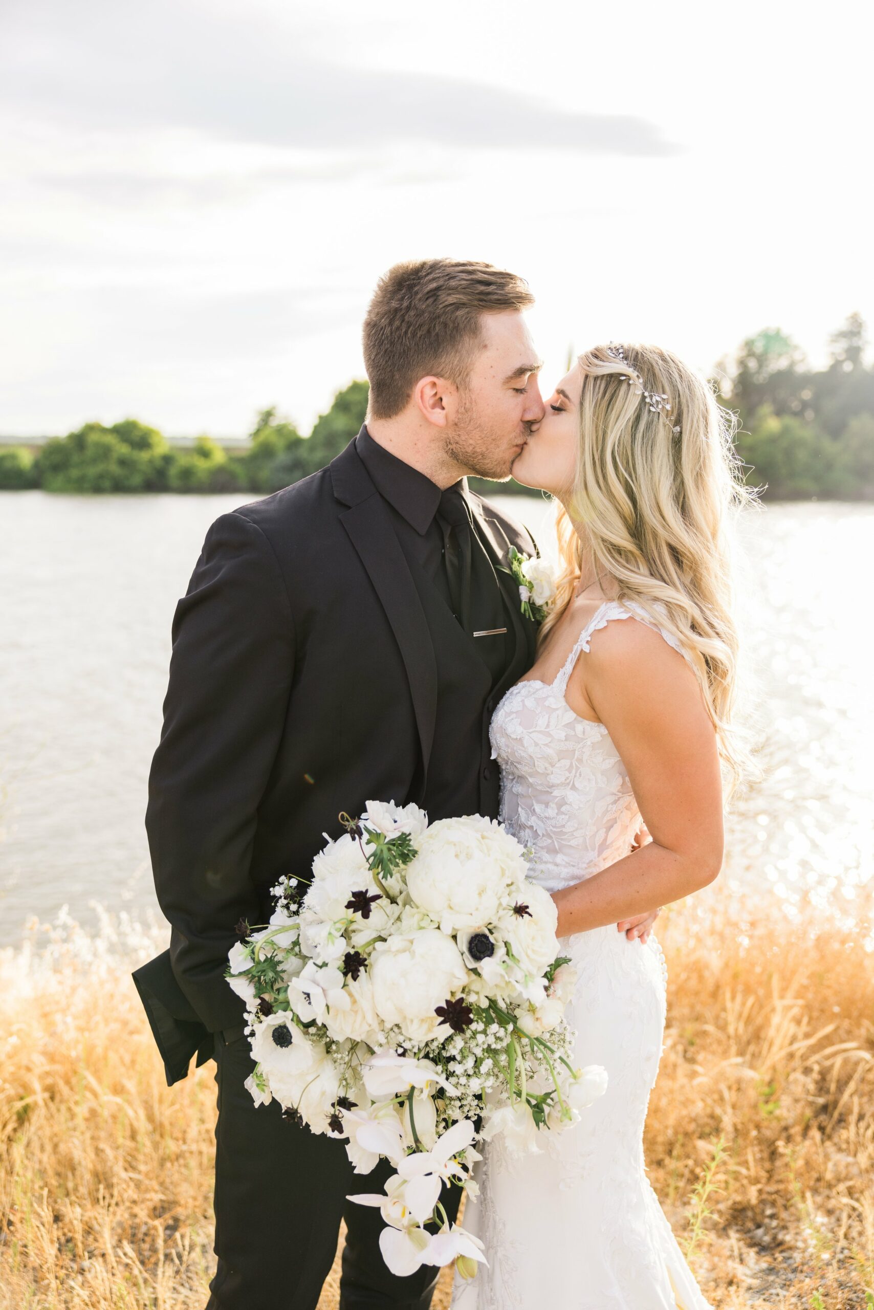 A couple dressed in formal wedding attire shares a kiss outdoors near a body of water; the bride holds a bouquet of white flowers.