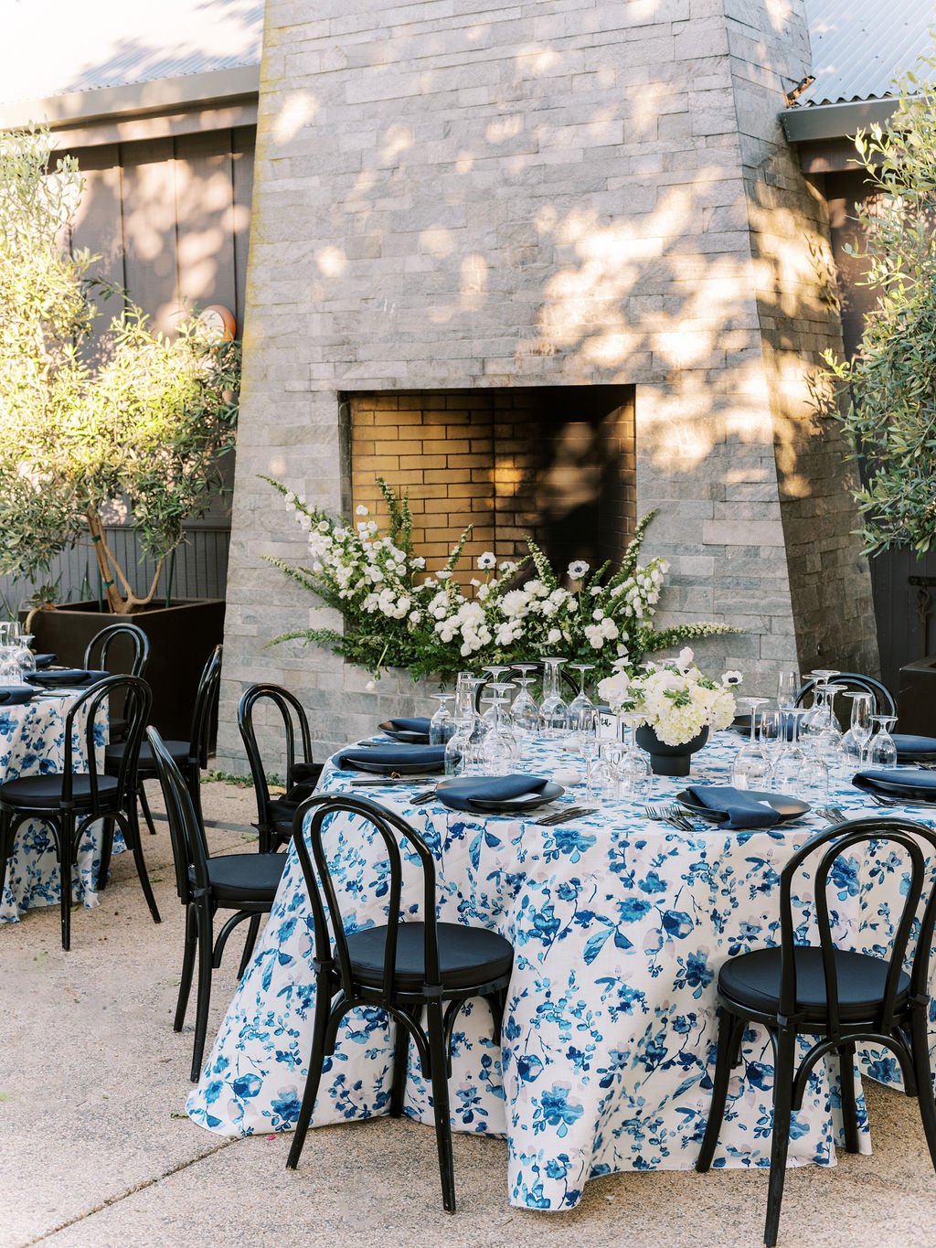 Outdoor dining area with tables covered in blue and white floral tablecloths, black chairs, and flower arrangements, set in front of a large stone fireplace.