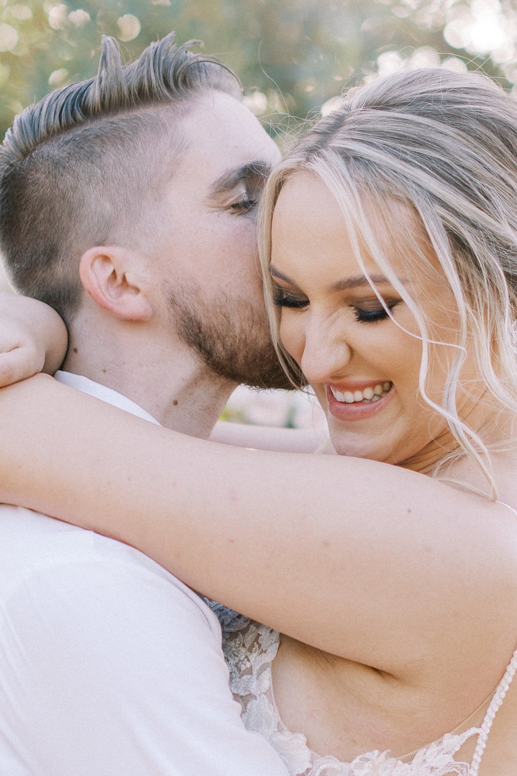 A man kisses a smiling woman on the cheek as they embrace, with greenery blurred in the background.