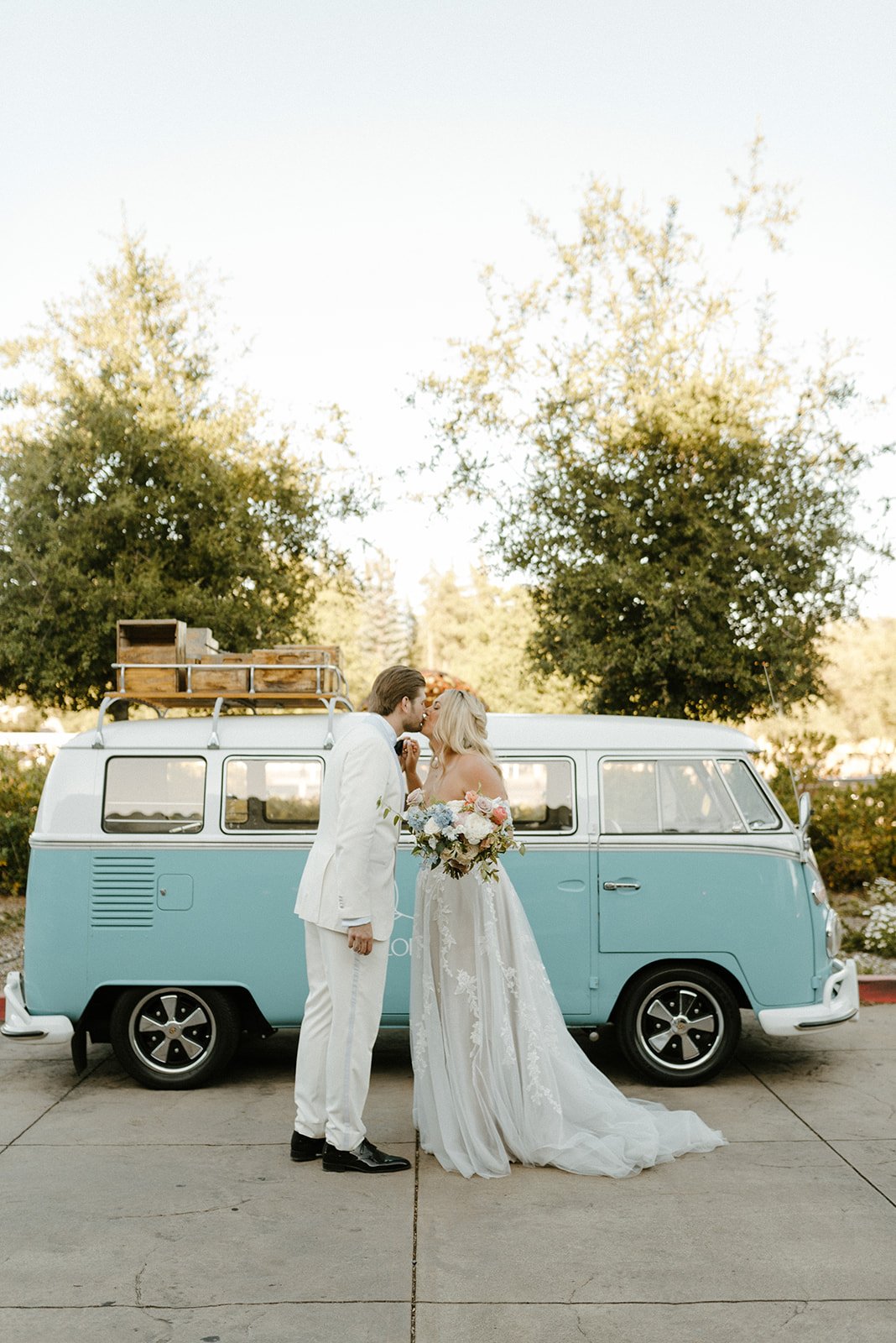 A couple dressed in wedding attire stands beside a vintage blue van. The groom, in a white suit, kisses the bride, who wears a white dress and holds a bouquet. Trees and clear skies are in the background.