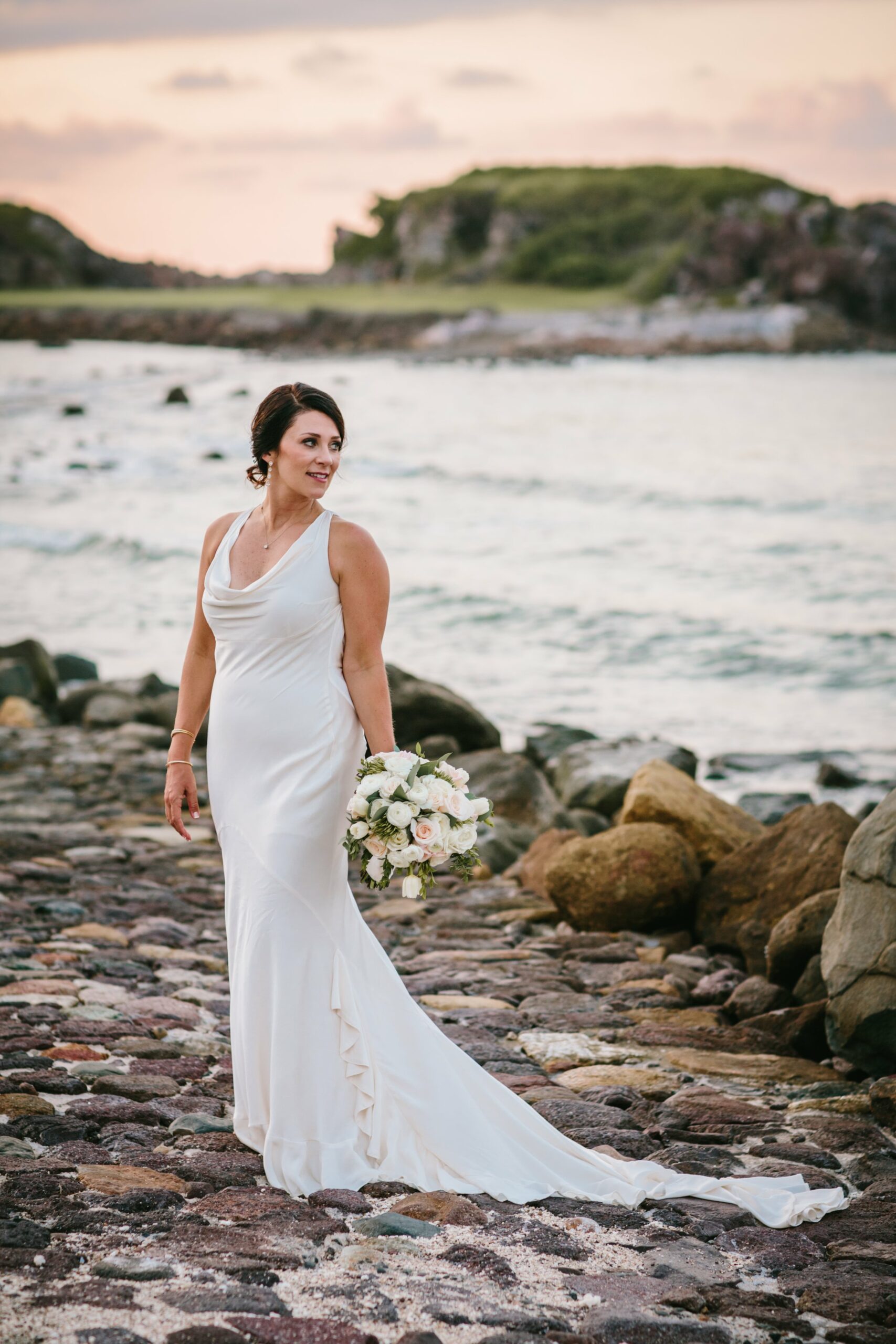 A woman in a white wedding dress stands holding a bouquet on a rocky shoreline with a distant view of the ocean and cliffs under a partly cloudy sky.