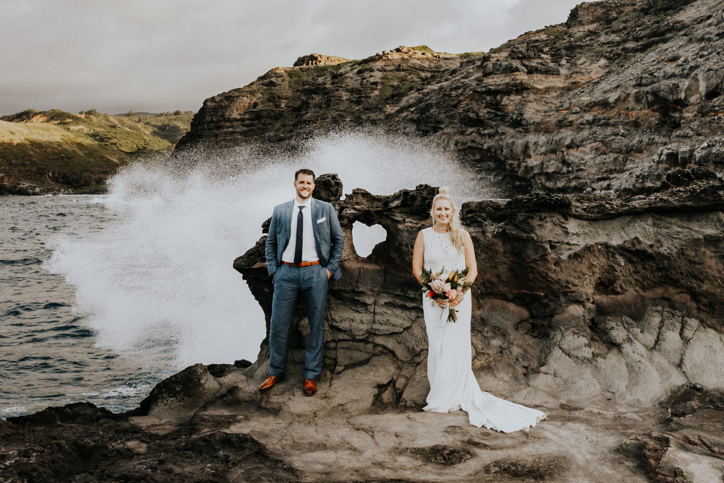 A couple in wedding attire stands on a rocky coastline with waves crashing behind them. The groom is in a blue suit, and the bride holds a bouquet.