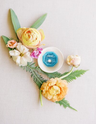A diamond ring in a blue box is placed on a small white dish surrounded by various colorful flowers and green leaves on a light beige surface.