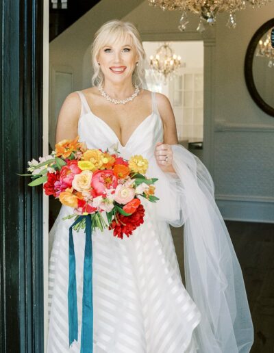 A woman in a white wedding dress holds a bouquet of colorful flowers. She is standing indoors near a chandelier, smiling at the camera.