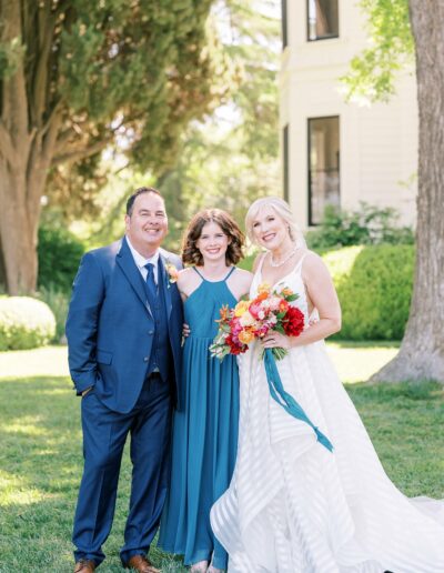 A man in a blue suit, a young girl in a blue dress, and a woman in a white dress holding a bouquet stand together smiling outdoors near a large house and trees.