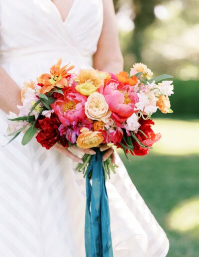 Person in a white dress holding a colorful bouquet featuring red, pink, orange, and yellow flowers with green foliage and a teal ribbon.