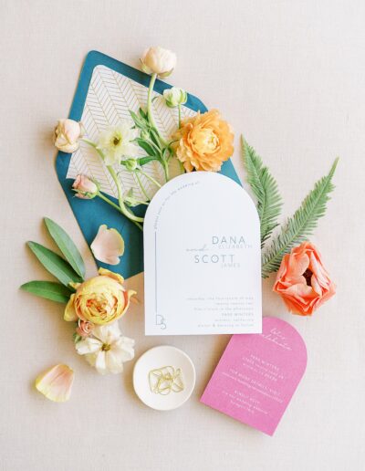 A wedding invitation set featuring a main invitation, RSVP card, and envelope, decorated with pastel flowers and greenery on a light background. Rings rest in a small dish beside the set.