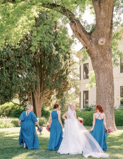 A bride and three bridesmaids, all in blue dresses, walk on a grassy area towards a large tree; sunlight filters through the leaves, and a white building is visible in the background.