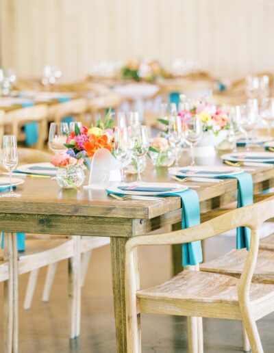 A wooden dining table is set with plates, glasses, and silverware, and decorated with colorful floral arrangements and teal napkins. Wooden chairs surround the table in a bright, airy room.