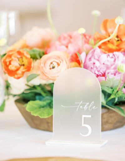 A table centerpiece with a frosted acrylic sign displaying "Table 5" in front of a floral arrangement featuring pink and orange flowers.