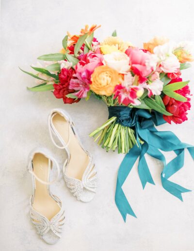 A colorful bouquet of flowers tied with a teal ribbon lies next to a pair of elegant, sparkling high-heeled shoes on a light surface.