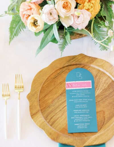 Wooden plate with a blue menu card on it, flanked by gold and white cutlery. Above the plate is a floral arrangement with various colorful flowers.