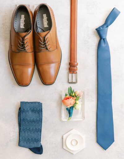 Brown dress shoes, a brown belt, a blue necktie, blue patterned socks, a floral boutonniere, and a hexagonal ring box are laid out on a light gray surface.