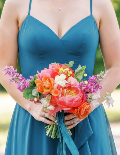 A woman in a teal dress holds a bouquet of vibrant flowers, including pink peonies and orange tulips, against a blurred outdoor background.