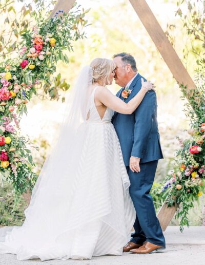A bride in a white dress and veil kisses a groom in a blue suit in front of a wooden arch decorated with flowers.