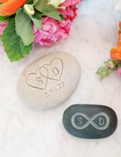 Two engraved stones; a light stone with hearts and "S&D" above "5.14.22," and a dark stone with an infinity symbol and "S&D." Bright flowers surround them on a marble surface.