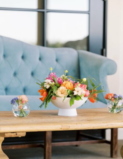 A white ceramic bowl of colorful flowers is centered on a wooden table, flanked by two small glass vases with flowers. A light blue tufted sofa and large window are in the background.
