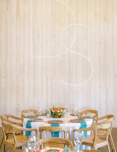 A round table with wooden chairs is set with teal napkins, wine glasses, and floral centerpieces against a white wooden paneled wall.
