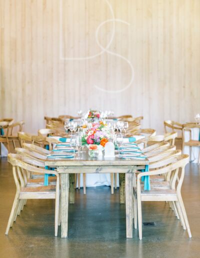 A rustic wooden table is set for dining with turquoise napkins, floral centerpieces, glassware, and wooden chairs in a light, airy room.