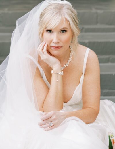 A seated bride in a white wedding dress and veil, resting her chin on her hand. She wears a pearl bracelet and a wedding ring.