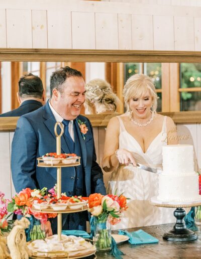 A couple stands together at their wedding reception. The bride, in a white dress, is cutting the wedding cake while the groom, in a blue suit, smiles. Flowers and desserts are on the table.