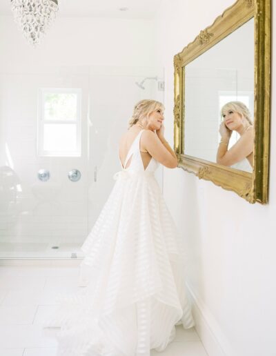 A woman in a white wedding dress looks at her reflection in a large, ornate mirror in a bright bathroom.
