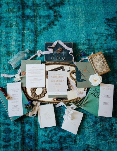 A collection of wedding stationery items, including invitations, menus, and envelopes, neatly arranged on a blue patterned surface, accompanied by decorative objects such as ribbons, a book, and a wax seal.