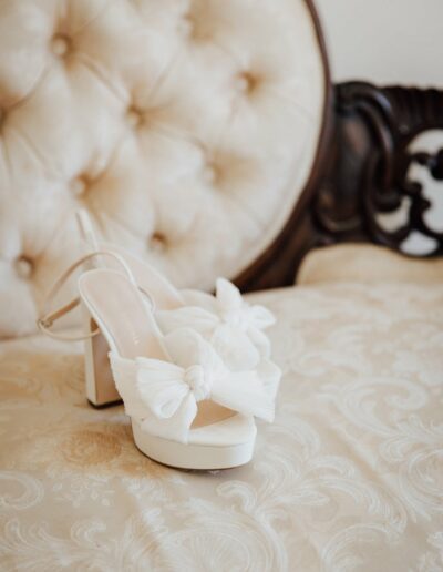 A pair of white women's high-heeled shoes with bow accents rests on a beige patterned cushioned chair with a tufted backrest.