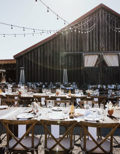 Outdoor wedding reception setup with long wooden tables and chairs in front of a rustic barn. Tables are set with plates, glasses, and centerpieces. Overhead string lights are strung across.