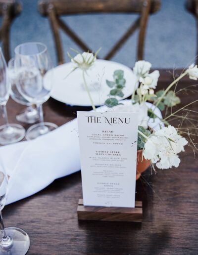 A wooden table set for a meal with wine glasses, plates, silverware, and a menu in a flower-filled vase at the center.