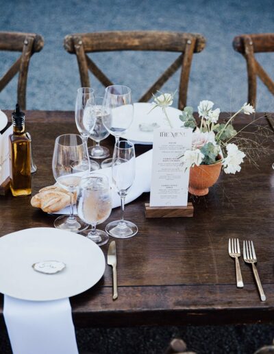 A neatly set wooden dining table with white plates, floral arrangements, menus, wine glasses, silverware, and a bottle of olive oil.
